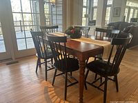 Six black wooden spindle-back chairs arranged around a wooden dining table in a dining room with hardwood floor and glass door in the background.