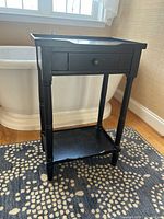 Front right angle view of the small black wooden side table showing drawer, shelf, and turned legs.