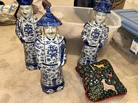 Three blue and white Asian porcelain figurines standing on table together, one small blue animal print pillow beside them.