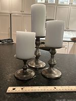 Silver-toned candlesticks with white candles, displayed with a ruler for size reference on a dark countertop.