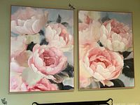 Front view of two floral canvas paintings in coordinating blush pink and green colors with wood framed edges hung on a green wall.