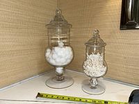 Two clear glass apothecary jars on a bathroom counter with a measuring tape showing approximate heights of 17 inches and 12 inches for the large and small jar respectively