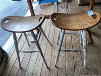 Two bar stools with wooden seats and white painted metal legs on wooden floor, showing wear.