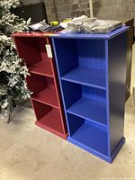 Front and side view of two 3-tier wooden bookcases, one blue and one red, showing their height and open shelves.