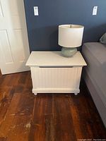 White lift lid chest next to bed with green ceramic lamp placed on top against dark wall