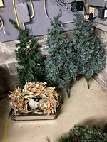 View of three artificial lighted Christmas trees with pinecone and berry decorations, standing on metal tripod legs in a basement corner with concrete wall and utility boxes in background. A fall-themed dried leaf and ghost decoration in a tray sits in front on the floor.