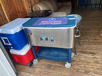 Three coolers and beverage buckets arranged together on wooden floor with couches and chairs in background.