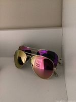 Two pairs of aviator style sunglasses, one with gold mirrored lenses and the other with purple mirrored lenses, shown stacked.