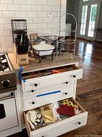 Wide view showing metal serving stands, knife block with set of knives, enamel cookware, baskets on counter, open drawers with towels, mitts, and utensils.