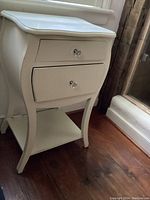 Front view of white nightstand showing two drawers with glass knobs, curved design, and lower shelf.
