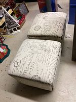 Two matching square ottomans with beige fabric featuring dark script text and postal stamp designs, decorated with metal nailhead trim on the bottom edges.