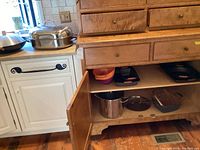 Wide shot showing multiple cookware items including pots, pans, colanders and roasting pans inside a wooden cabinet and on countertop.