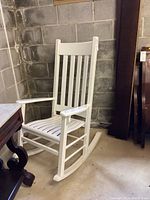 White wooden porch rocking chair standing on concrete floor by a concrete block wall in basement, showing full chair profile from left side.
