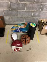 Photo of all items together including Indo Board balance trainer, black roller, and sports balls on concrete floor against cinder block wall.