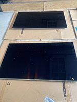 Two Samsung 55-inch flat screen TVs laid face up on cardboard showing their black screens with some dust.