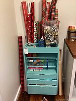 Photo of turquoise wrapping supplies cart holding various patterned rolls of wrapping paper and bows on top.
