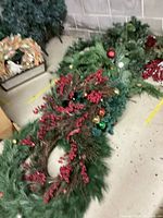 Overall view of green artificial garland decorated with red berries and multi-colored ornaments on the floor near gray brick wall.