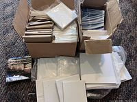 Photo showing two open cardboard boxes filled with multiple stacks of white square tiles mostly wrapped in protective paper, with some loose tiles around including printed tiles with nature scenes.