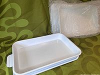Photo showing two white rectangular kitchen pans nested inside two matching trays, glossy finish, placed on patterned green fabric with bubble wrap in background.