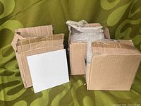 Two open cardboard boxes containing frosted glass tiles wrapped in bubble wrap, showing two glass tiles outside the boxes placed on fabric background.