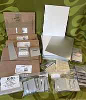 Photo showing stacked white gloss aluminum panels with cardboard backing and boxes containing various metal wall mounts and easel stands, all placed on a green fabric background.