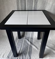 Front view of black square end table with four removable white tiles on top placed on a grey fabric background.