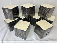 Stacks of white sublimation coasters shrink wrapped in plastic, showing the white printed top and the dark cork or MDF back layer, arranged in multiple piles on a white surface.