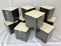 Multiple stacks of square white coasters with cork backing shrink-wrapped in plastic, arranged on a light surface.