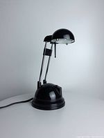 Front angle showing black halogen desk lamp with dome-shaped shade, telescoping arms, and round base on white surface.