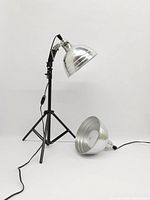 Two aluminum cone-shaped reflector photographer's lights, one mounted on a black tripod stand with adapter, cable and switch visible, shown against white background.