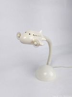 Side view of one white ceramic airplane table lamp with flexible neck and dome base on a plain background.