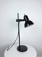 Black adjustable desk lamp with round base and vertical pole, shown against white background, lamp not lit. Shows lamp's design and condition.