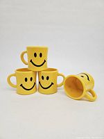 Photo showing three yellow plastic smiley face mugs stacked with one mug laying on its side, displaying the hollow interior and handle design.