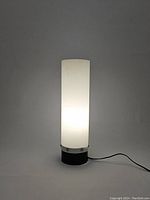 Straight-on view of the vintage lamp illuminated showing the tall white cylindrical tube and black base with power cord.