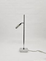 Full view of the lamp, showing vertical stand, squared base, and the cylindrical slotted lampshade angled forward.