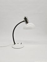 Desk lamp with white half dome shade, chrome perforated shade connector, flexible gooseneck, round white metal base with two control knobs and power cord visible.