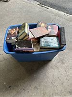 Blue plastic bin filled with a variety of books including hardcover and paperback editions, viewed from above at an angle showing book covers and edges.