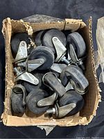 Lot of 15 swivel casters with metal brackets and black rubber wheels inside a worn cardboard box and outside it on a black background.