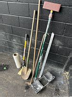 Photo showing nine construction hand tools arranged against a wall including shovels, tamper, measuring ruler, material roll, and caster wheels.