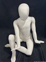 White child mannequin posed sitting on a black surface with one knee bent and one arm resting on the knee.