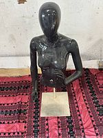 Black glossy half body female mannequin on square metal stand with metal rod support, photographed front view on patterned red and black fabric surface