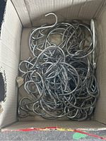 Top view of a cardboard box filled with over 50 metal S-shaped display hooks, silver color, piled on top of each other.