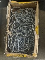 View of a worn cardboard box filled with over 50 shiny chrome-finish metal display hooks piled tightly. Hooks have round bends for hanging.