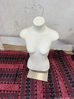 White female torso mannequin with no head or arms, mounted on a rectangular metal base standing on a patterned fabric surface.