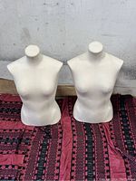Two female torso mannequins photographed from the front on a red patterned cloth, showing the full torso shape with no arms or heads, cream/off-white fabric covered.