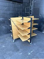 Dual-sided wooden display shelving unit with four wooden shelves on each side and black metal frame, shown outdoors next to black brick wall. Four caster wheels under frame.