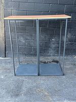 Front view of two display mannequin stands side by side showing black metal frames, square bases, and wood tops against a gray brick wall background.