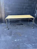 Front view of the rectangular display table on metal legs and caster wheels with wooden top, set against a painted brick wall.