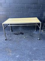 Front view of rectangular display work table showing wooden top and metal frame with caster wheels.