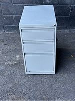 Front view of white 3 drawer metal filing cabinet with wheels, showing lock without key.